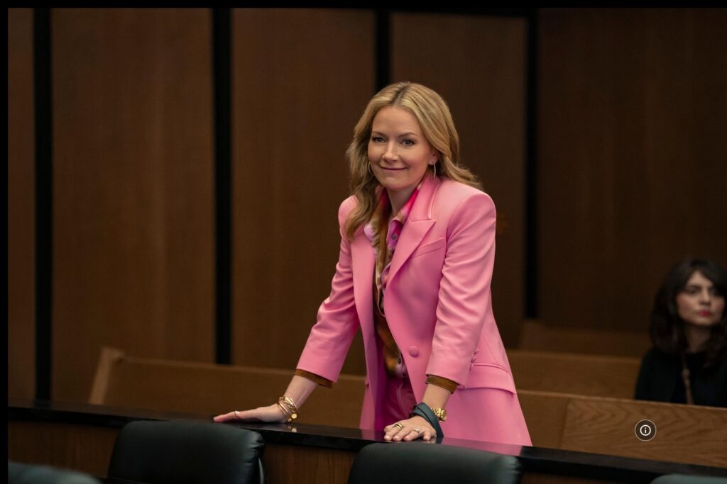 Lorna Crane from The Lincoln Lawyer, wearing pink jacket and leaning on a courtroom table.